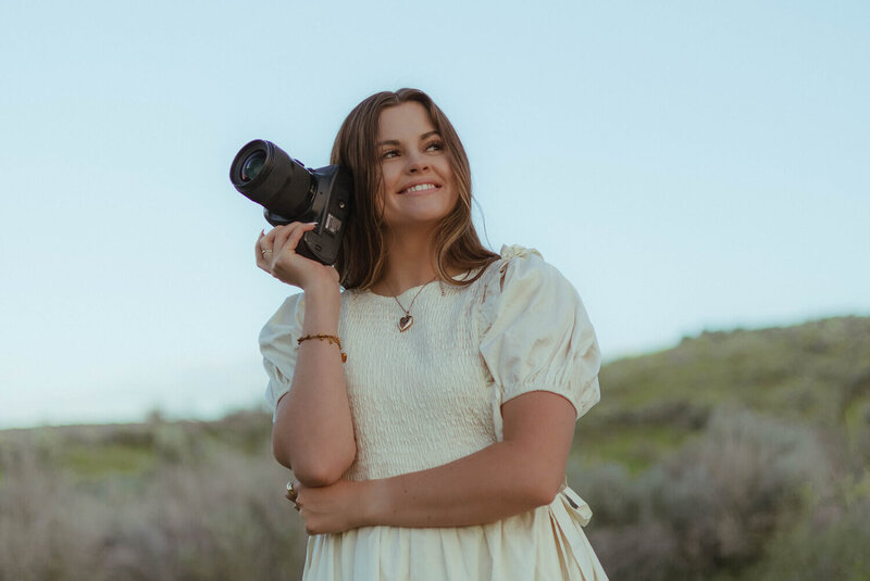 Standing on a hill, Brittney looks off in the distance while holding her camera to her head.