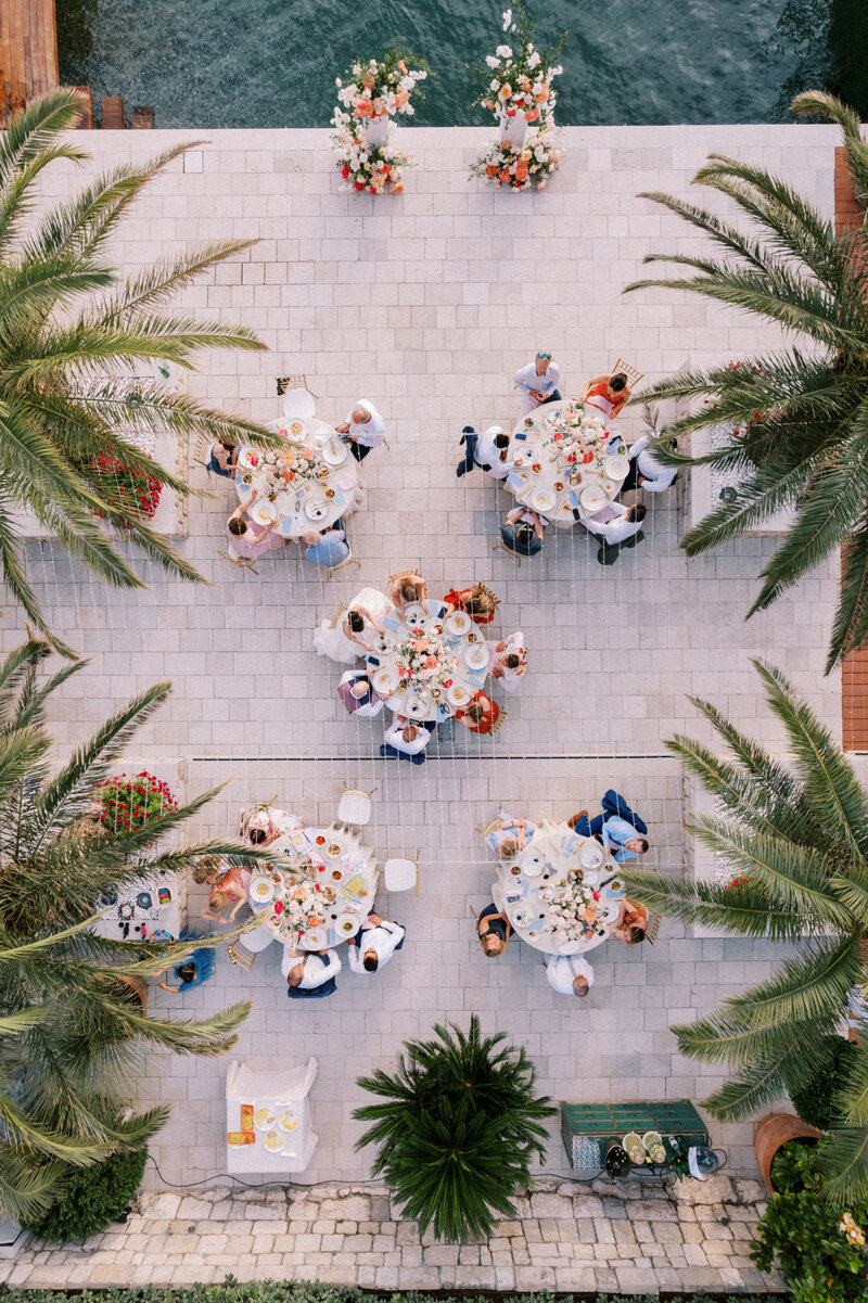 Aerial view of elegant outdoor wedding reception by the sea with floral arrangements, round tables, and palm trees, captured by a fine art wedding photographer
