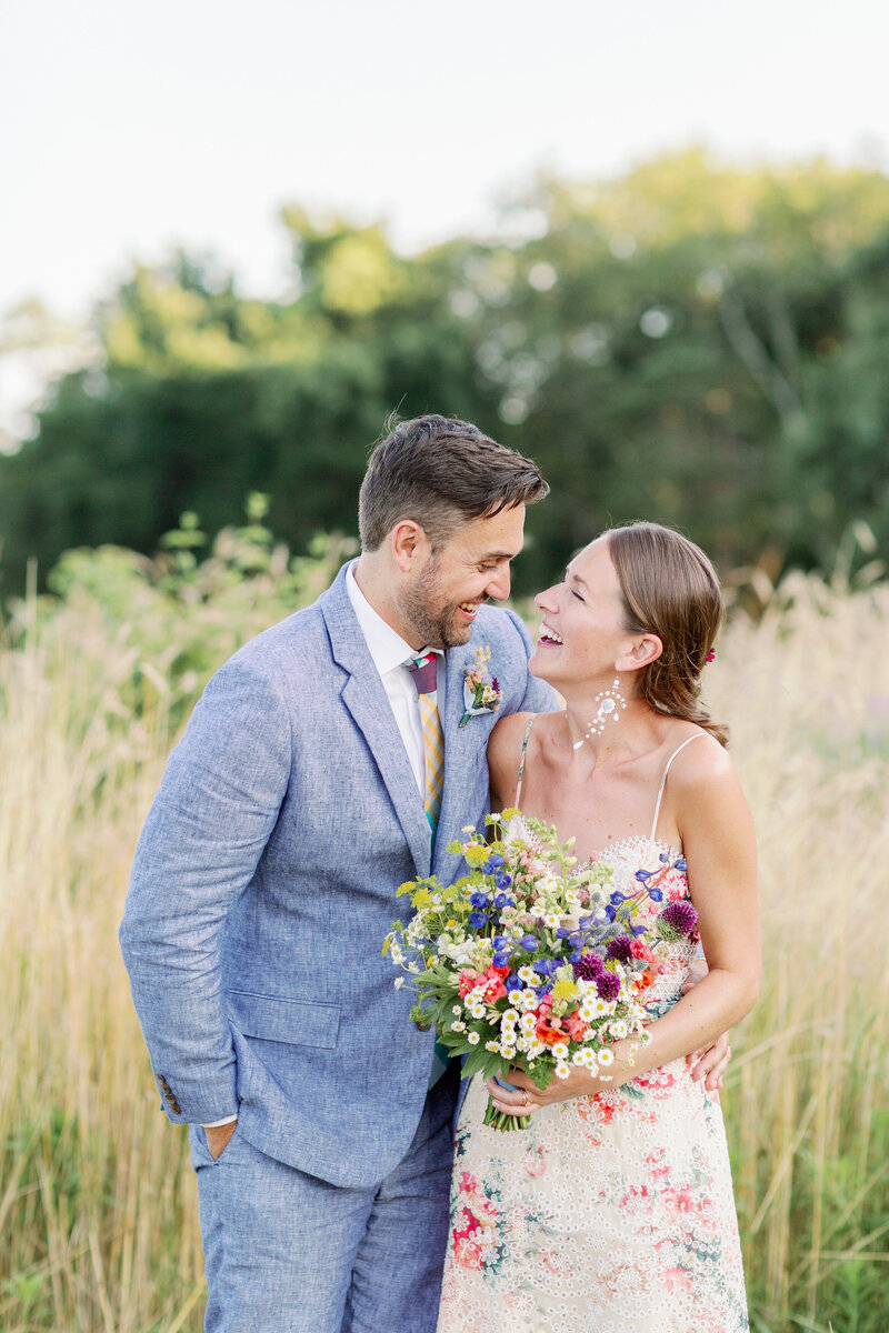 Couple standing in a large field smiling at each other as one holds a bouquet of flowers.
