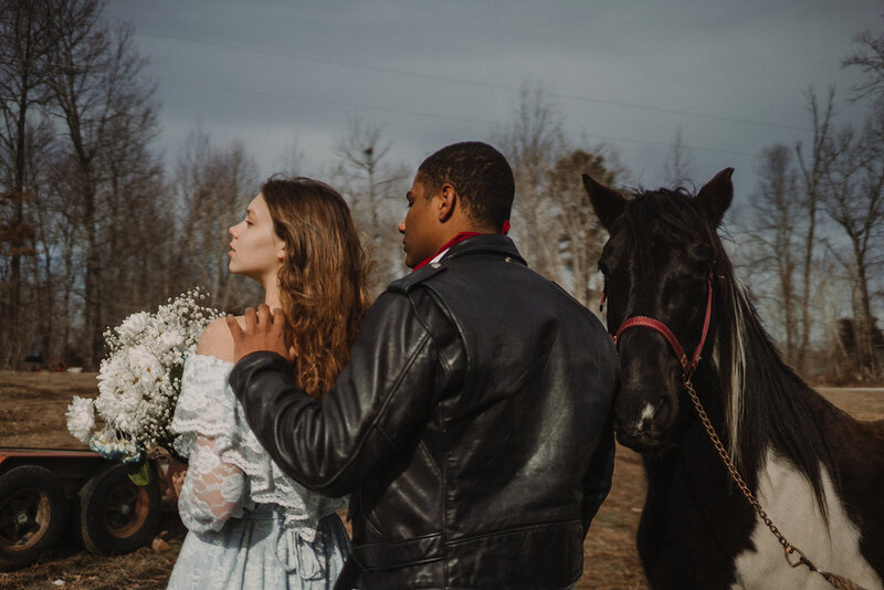 Alternative elopement couple in a leather jacket and vintage wedding dress pose with a horse and daisy bouquet.