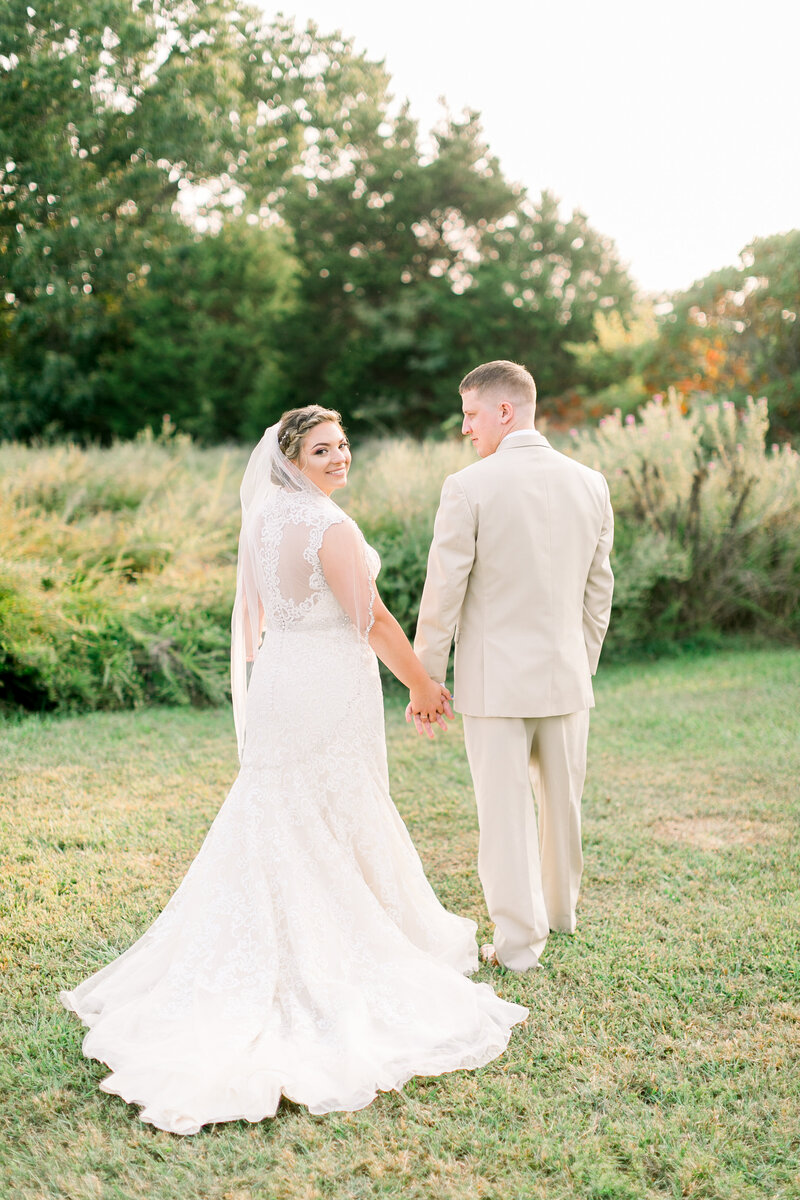 bride-and-groom-walking-and-holding-hands-groom-wearing-khaki-tux-bride-wearing-elegant-lace-wedding-dress-and-mid-length-wedding-veil-greenhouse-two-rivers-wedding-in-springfield-mo-by branson-wedding-photographer-kathryn-faye-photography