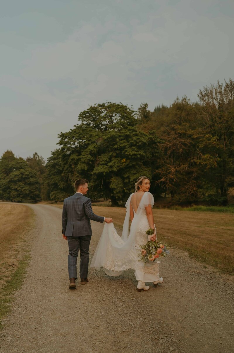 A bride and groom walking hand-in-hand on a gravel path surrounded by lush greenery. The bride wears a lace wedding gown with flutter sleeves, holding her bouquet and her train, which the groom gently assists with. The groom is dressed in a checkered suit, and they are framed by trees and an open field under a soft, overcast sky.