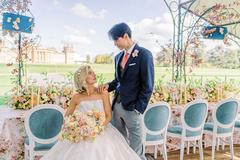 a groom stands next to his bride who sits in a teal chair at a long wedding dinner table that is decorated with pink linen and yellow and pink flowers at blenheim palace