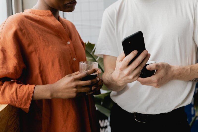 Close-up of two people, one holding a coffee cup and the other a smartphone, engaged in a conversation. The setting includes casual attire, plants in the background, and a relaxed atmosphere.