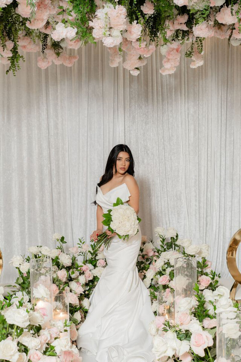 Bride in white gown holding bouquet, posing in floral ceremony setup