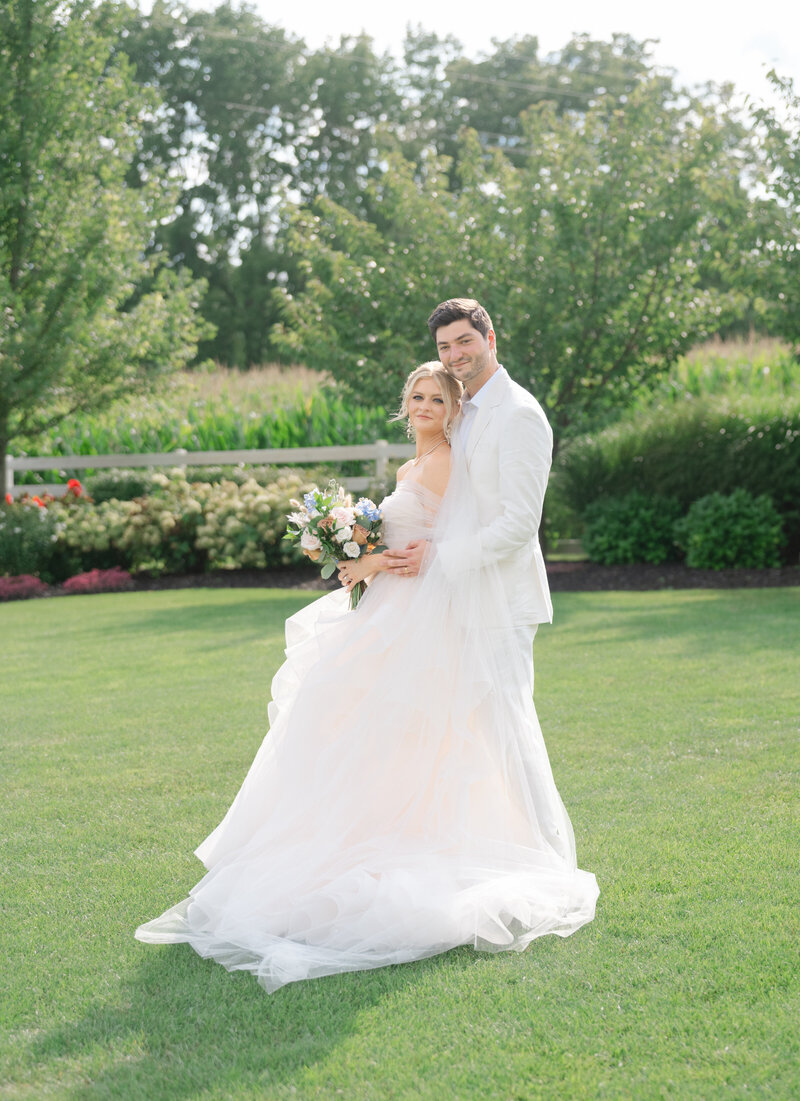 A bride and groom in a white suite pose in a garden at their wedding in France