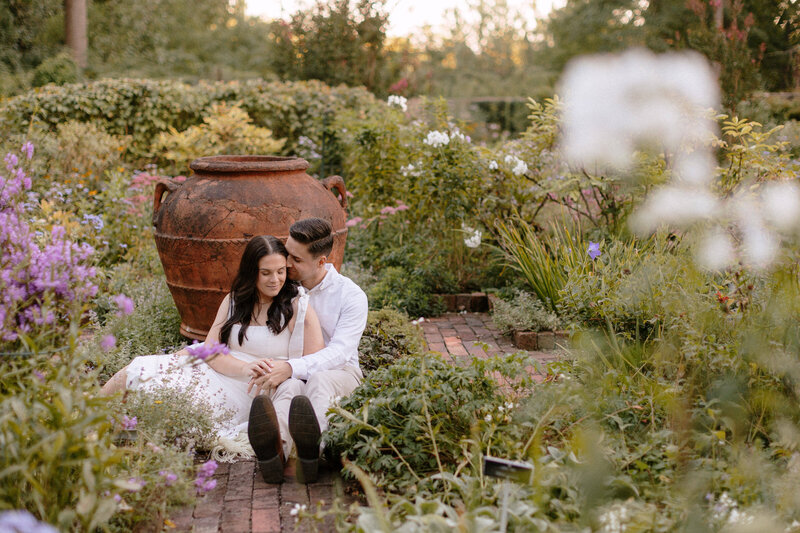 A couple sits together on a brick path, surrounded by lush greenery and colorful flowers, with a large clay pot behind them.