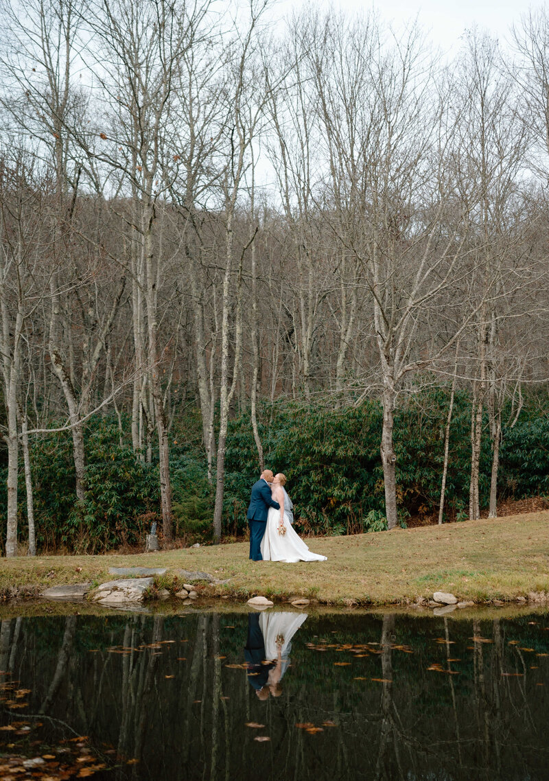 The bride and groom share a kiss in a beautiful setting surrounded by a lake and trees.