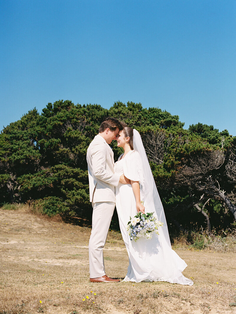 A bride and groom with their foreheads resting against each other with a clear blue sky, representing Heather's wedding portfolio. 