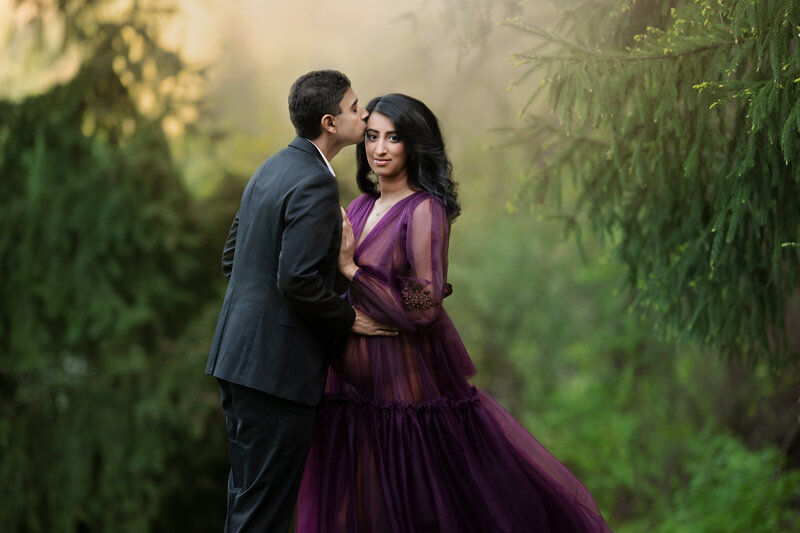 Couple with pregnant woman standing in front of pine trees.  She is wearing a purple sheer gown and he is wearing a dark gray suit.
