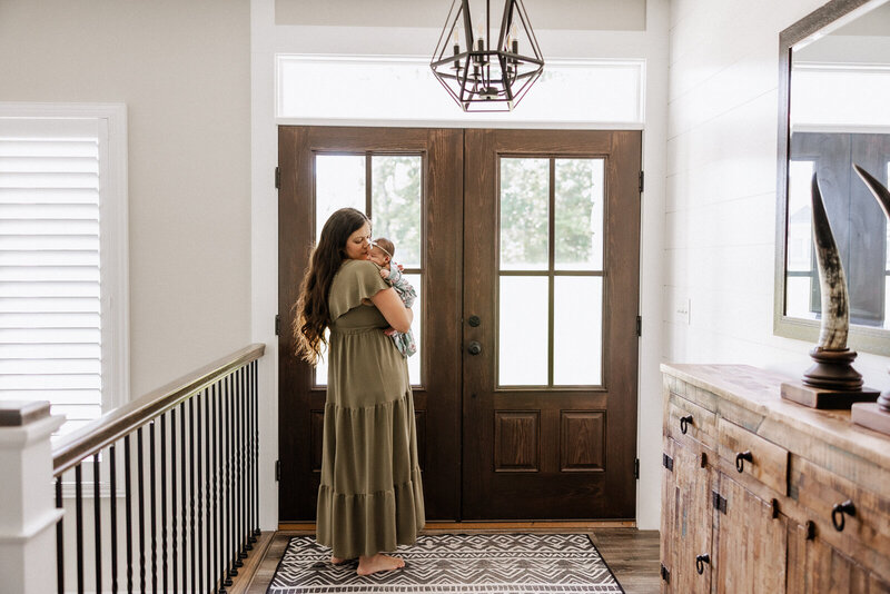 In-home lifestyle newborn session portrait of a mother standing in her foyer in front of the door while holding and rocking her baby in Festus, Missouri. 