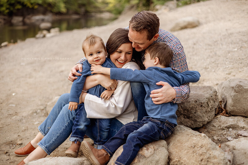 Smiling family of four sitting on rocks near a riverbank, sharing a warm embrace during an outdoor family photography session in Grand Rapids, Michigan