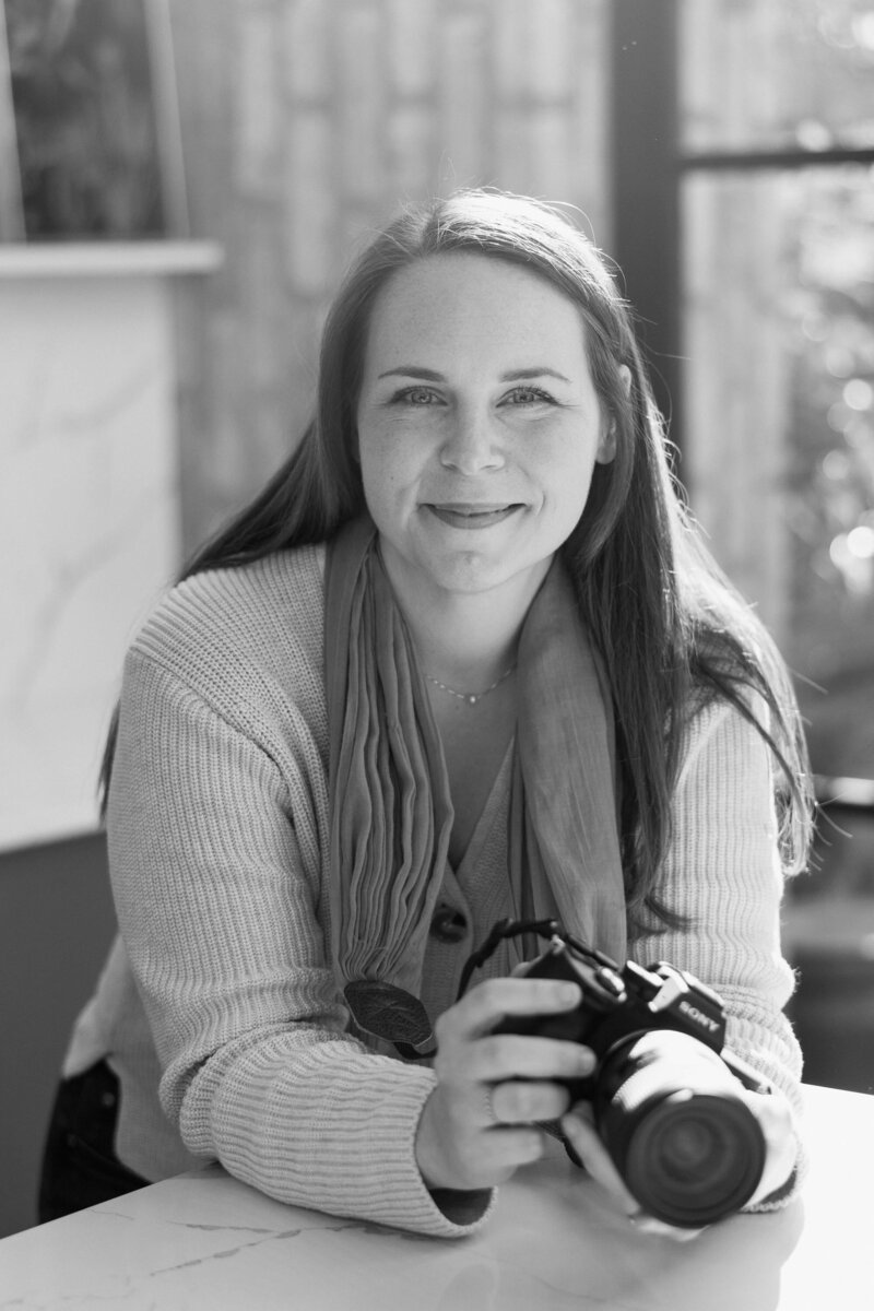 A Munich photographer wearing a cream sweater and coral scarf smiles while holding a camera in a bright sunlit room.