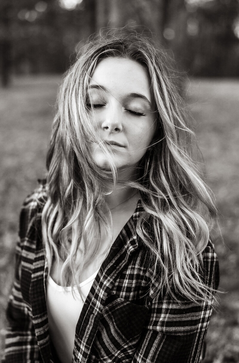 Black and white close up portrait of a senior girl closing her eyes while the wind gently blows her hair across her face. 