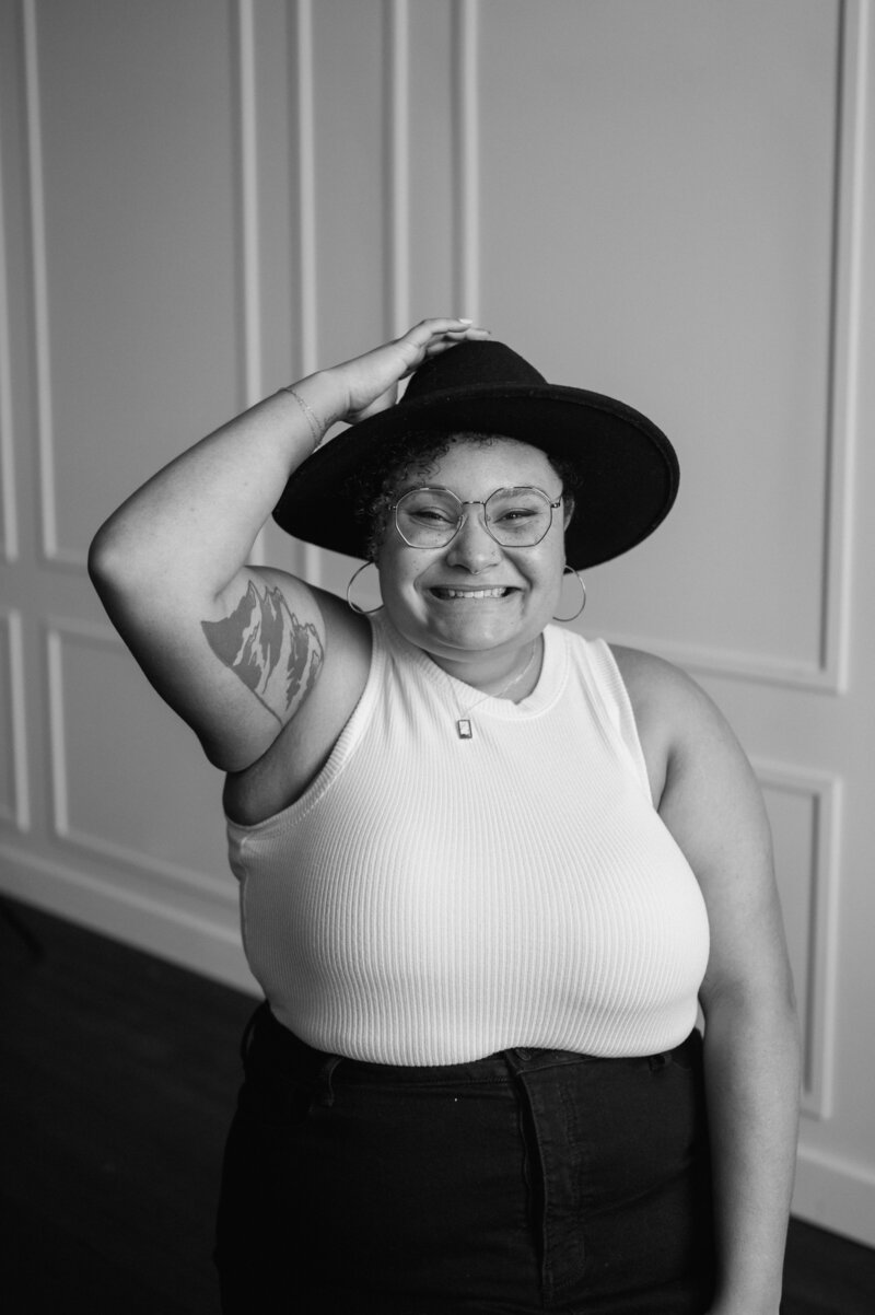 Ashley, an elopement photographer based in the PNW with a mountain tattoo, wearing a black hat, white tank top and black jean skirt. She's wearing the coolest gold wired frame glasses and has a massive smile on her face. 