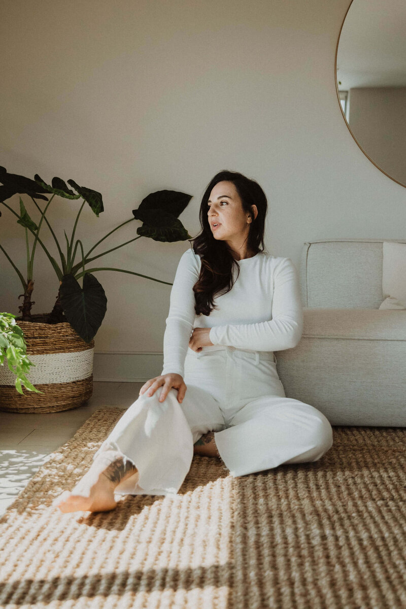 Holistic Esthetician, Jen Stoeckert, wearing a white top and white pants sitting on a jute rug leaning against a neutral colored couch