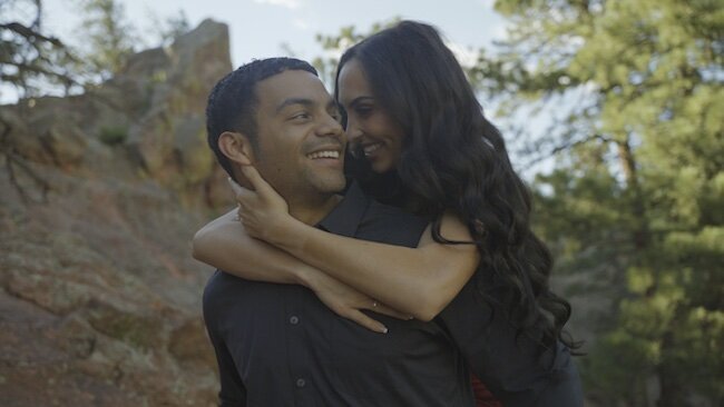 A man stands behind and wraps his arms around a woman in a wedding dress. There is a rainbow flare.