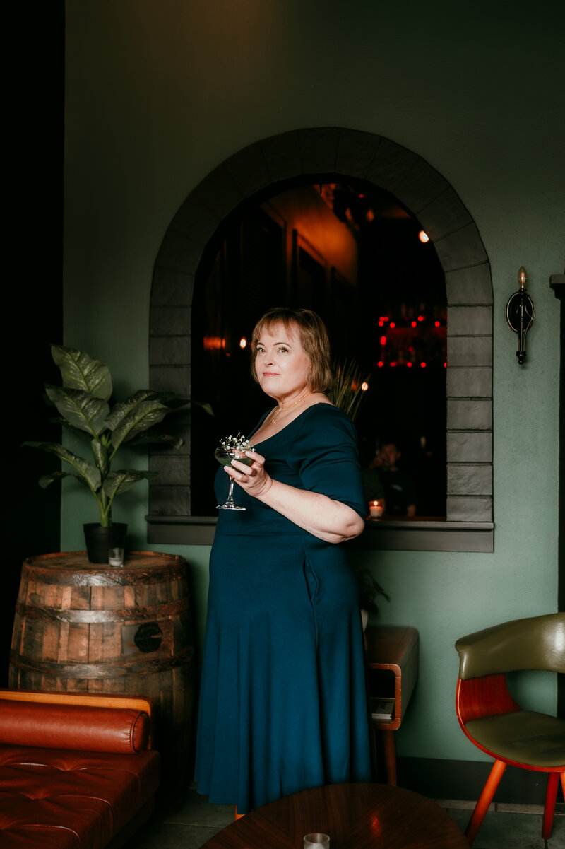Confident woman in a teal dress holding a cocktail during a Minneapolis online dating photo session at a stylish Bar Cart lounge