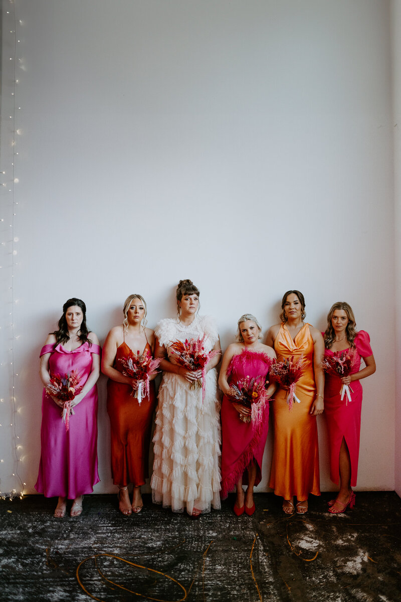 A bride and her bridesmaids who are wearing a combination of pink and orange dress stand in front of a white wall at Trafalgar Warehouse in Sheffield. 