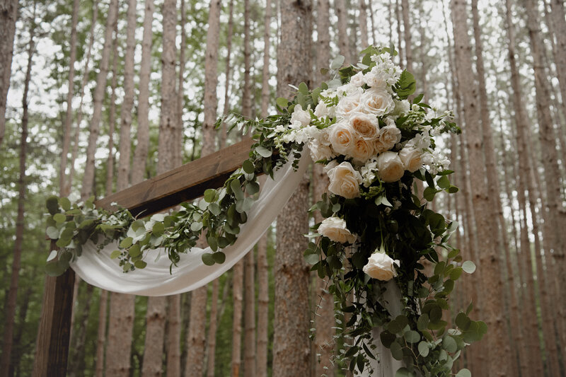 A rustic dark wood wedding arbor draped with white fabric, greenery, and white flowers stands in a pine grove in upstate New York