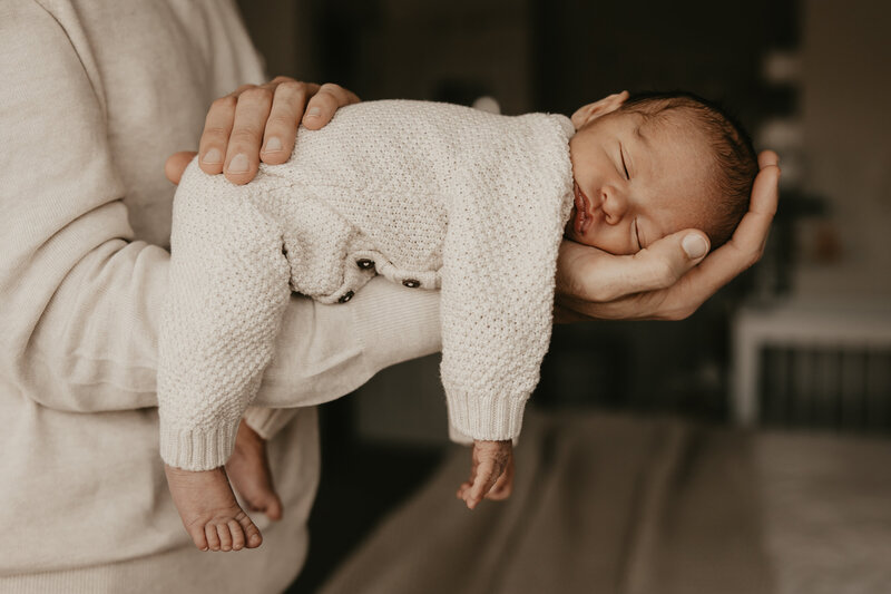 Newborn baby sleeping in a cozy knit outfit, gently cradled in a parent’s arms during a lifestyle newborn session in Metuchen, NJ, captured by Burkat Photography.