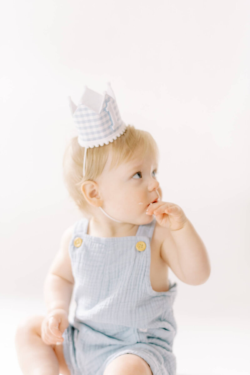 A one year old baby boy wears a birthday crown and licks frosting off his finger while tasting his first cake.