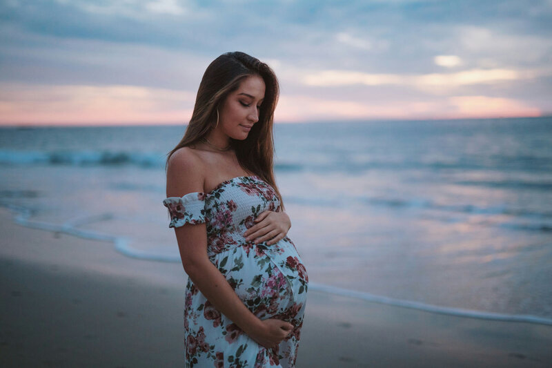Pregnant woman in a floral dress is standing on the beach holding her belly