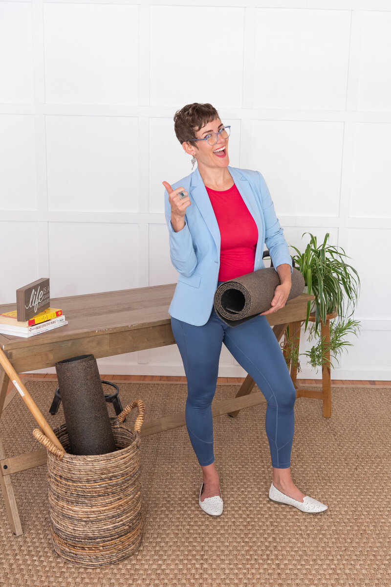 A woman in yoga attire holding a yoga mat and smiling with joy.
