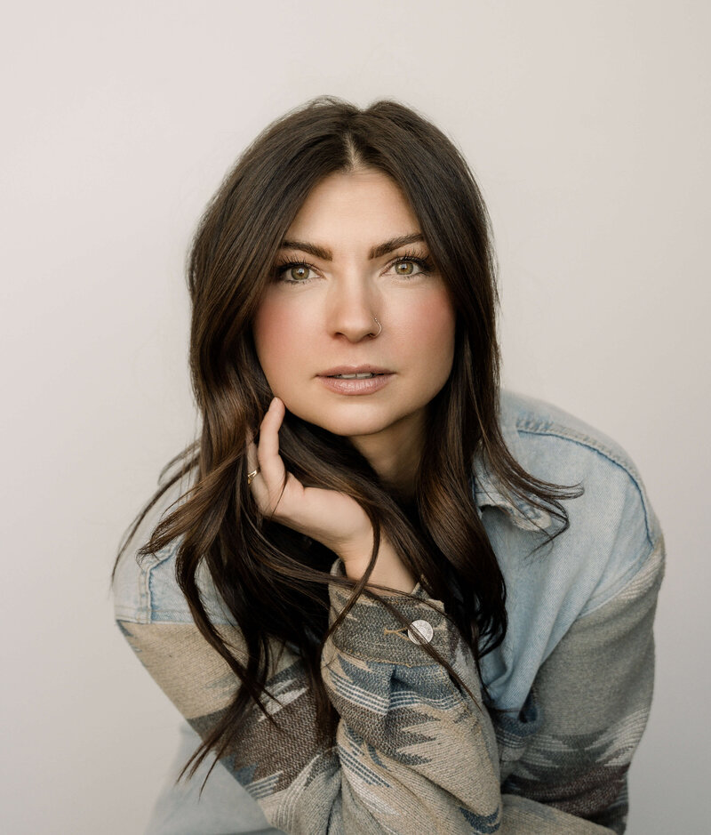 A long haired brunette photographer in Denver smiles while making eye contact .  She is dressed in denim jacket with a western aztec pattern.