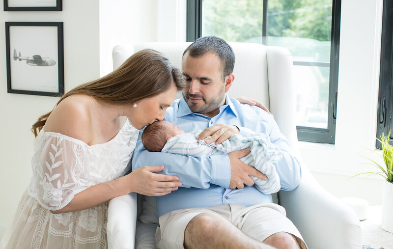 New dad sitting in the white chair holding his newborn son and mom is kneeling next to them and kissing the baby on his head.  Beautiful picture of the purest love with a newborn baby. 
