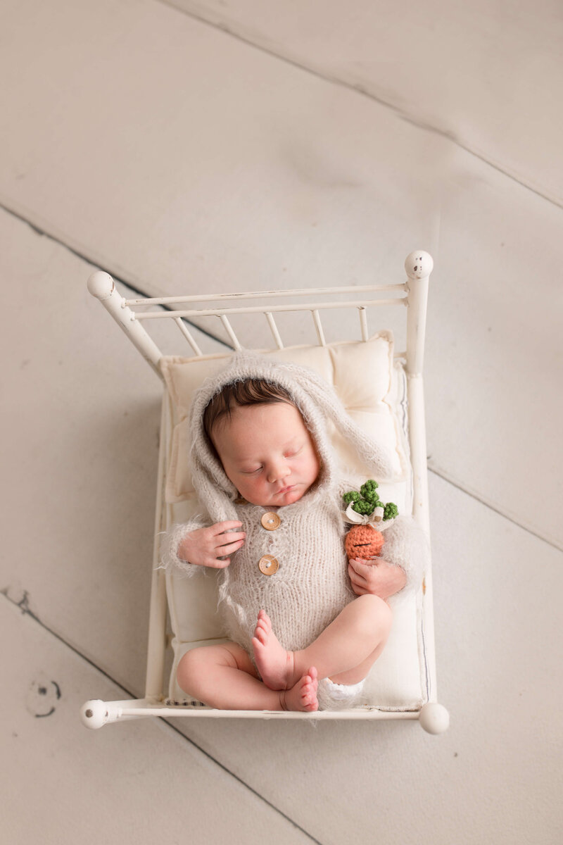 Infant girl wearing bunny outfit holding a knit carrot in a bed