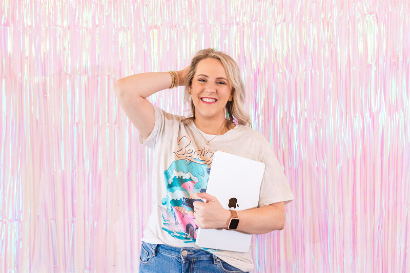 A person smiling and holding a closed laptop while standing against a pink metallic fringe background, celebrating her VIP launch day with an elegant Showit website template designed for women entrepreneurs.