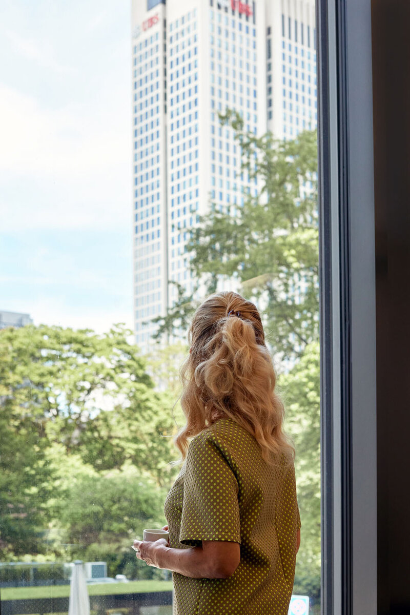 Roxana back to the camera, holding a coffee cup looking through a window to the Frankfurt skyscrapers