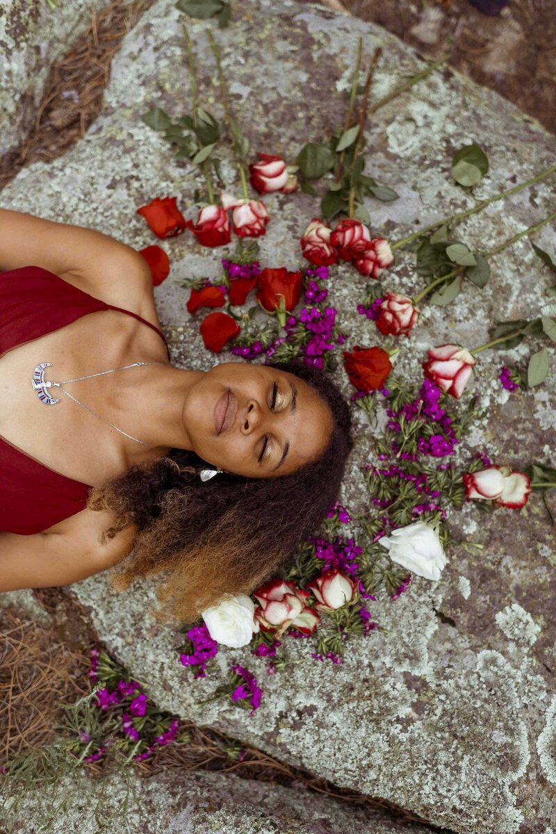 woman lying on a rock with her eyes closed and red, white and purple flowers all around her head