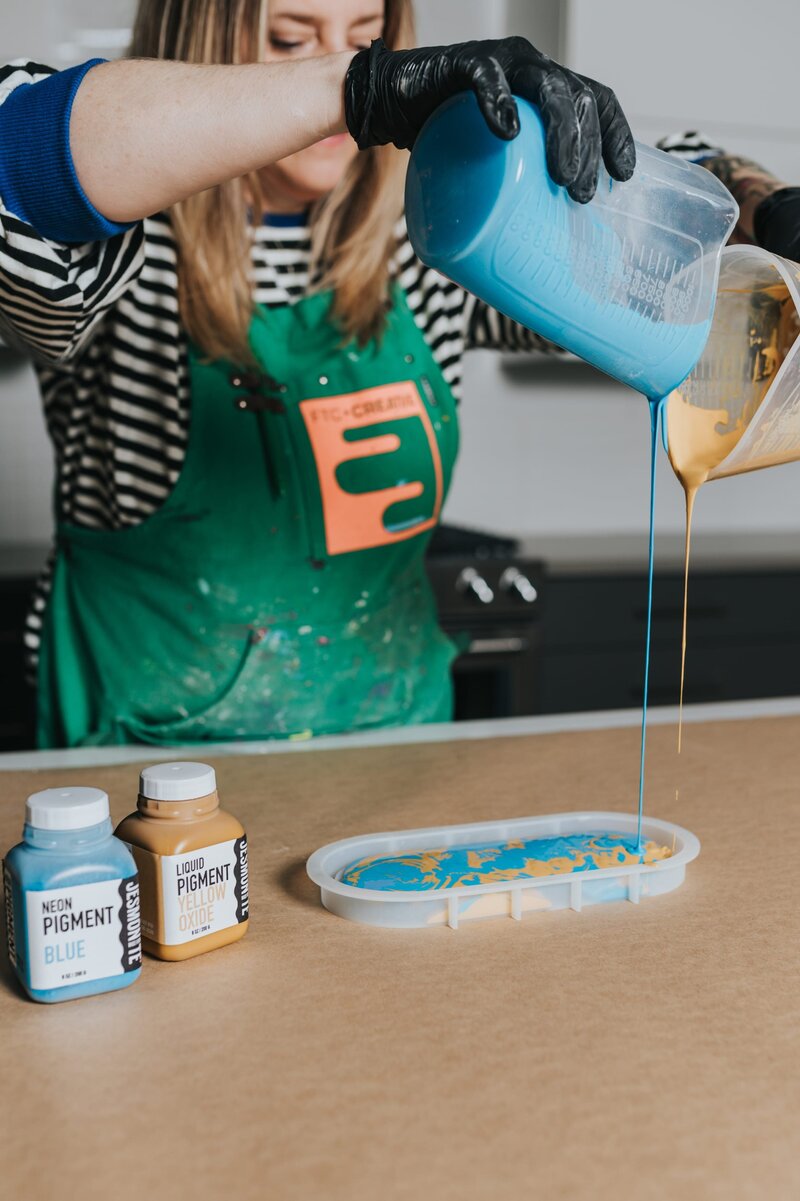 woman in green apron pours blue and yellow paint into a tray.