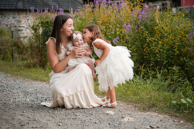 A mom at Boston Hill Farm crouches with her baby and smiles while her toddler kisses her baby's cheek.