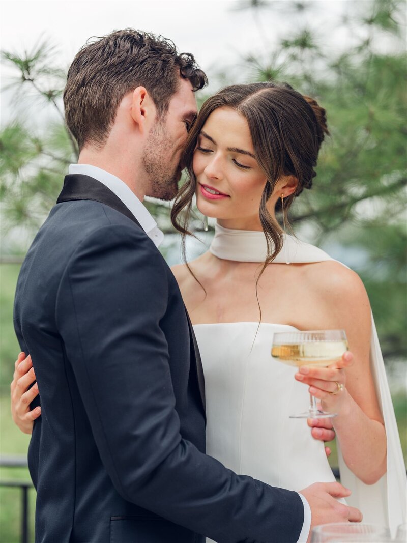 Wedding portrait of a married couple in front of an orange helicopter during their elopement. the bride is wearing a white strapless wedding dress and holding a bouquet or green and white flowers, the groom is wearing a black suit with white shirt looking at the bride's shoulder