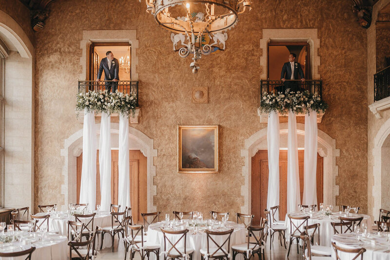 Two men stand on small balconies above a dining area with round tables set for an event. The room features ornate decor with floral arrangements, white drapes, and a chandelier—an elegant setting perfect for a Banff Alberta wedding.