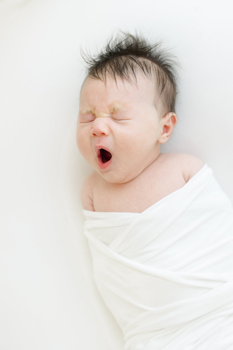 Swaddled newborn baby boy yawns during newborn photography session