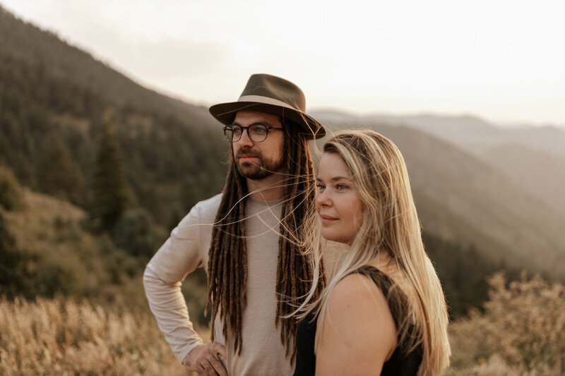 Man with dreads, glasses, and a wide brimmed hatand woman stand on foothills of mountain looking off to the distance as woman's blonde hair blows in the wind.