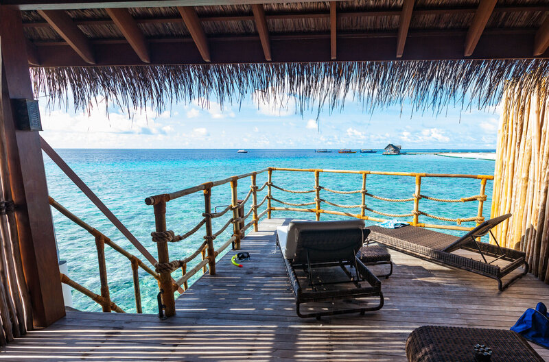 A wooden deck overlooking crystal clear blue ocean waters, with two lounge chairs and a tropical thatched roof. The view includes a small boat and an island in the distance under a partly cloudy sky.