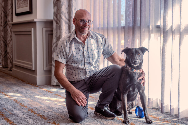 Man wearing glasses and a patterned shirt kneeling beside his dog in a softly lit indoor setting, photographed by Vyrl Photo, showcasing Tucson brand photography.