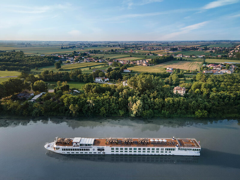 A long white river cruise ship traveling along a calm river, bordered by dense green trees, with expansive farmlands and scattered houses stretching into the distance under a clear blue sky.
