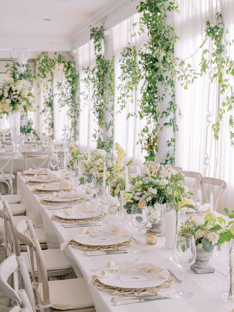 Elegant wedding table setup with white linens, gold accents, and greenery along the windows.