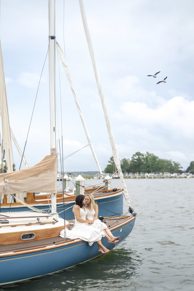 Two affectionate women in white dresses sit together on the edge of a blue sailboat docked in a marina. Their feet dangle over the water. The sky is partly cloudy, and a few birds fly above. In the background, there are trees and buildings near the shore.