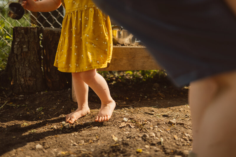 detail of toddler girl feet walking barefoot on dirt in mud kitchen in the backyard while wearing cute yellow summer sundress