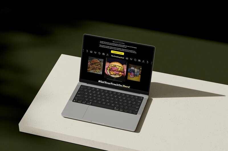 A laptop displaying the Patty Freaks website, highlighting their menu with images of stacked burgers and beverages. The screen features a header with the text "Get Your Freak On Here!" and details about their food offerings. The laptop is placed on a light-coloured surface against a dark background, emphasising the vibrant and appetising visuals on the screen