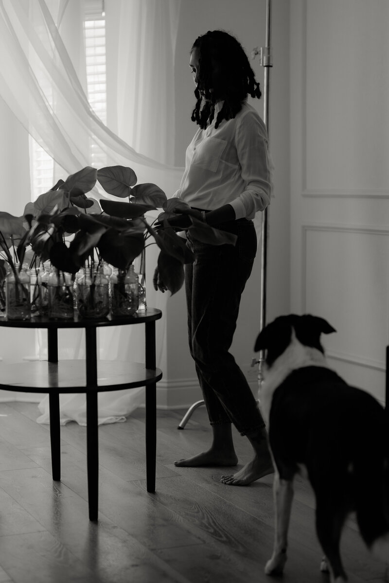 soulful black and white photo of a Black woman maternity photographer from the DFW area, standing in her Fort Worth studio, admiring her plants on a table, with her dog nearby, capturing a serene and personal moment