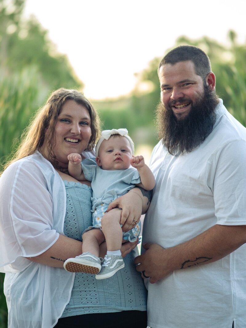 mom and dad smile and hold their baby girl together for an outdoor family portrait session at Fountain Creek Park in Colorado. they wear white and light blue.