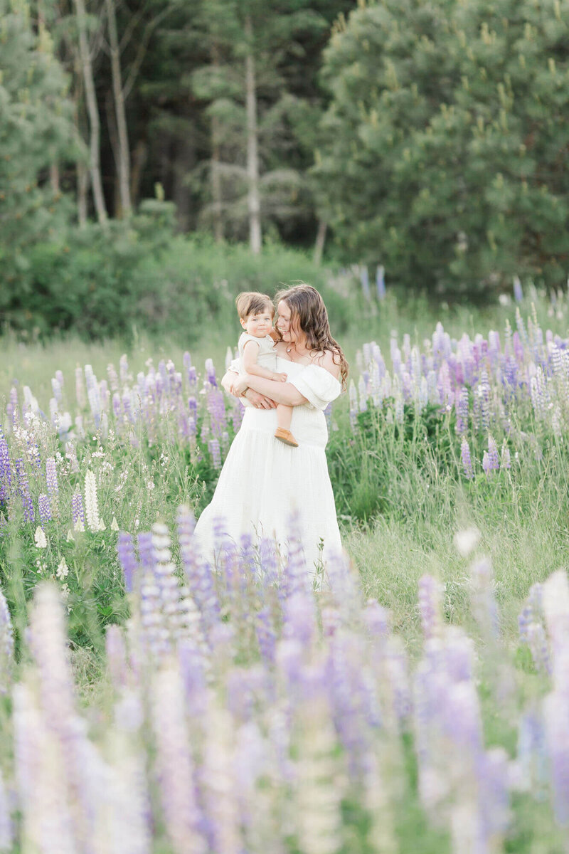 Photograph of Photographer Ashlie Behm with her 1 year old baby out in a field of wild lupine flowers. They are both wearing light, neutral tones and colors. 